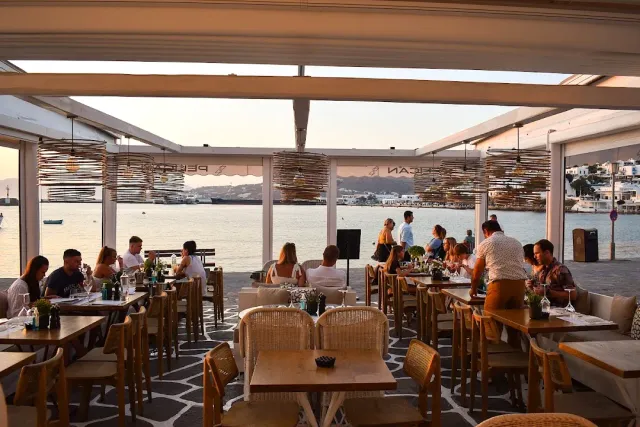 A covered seafront terrace with wicker lamps and diners at wooden tables at dusk