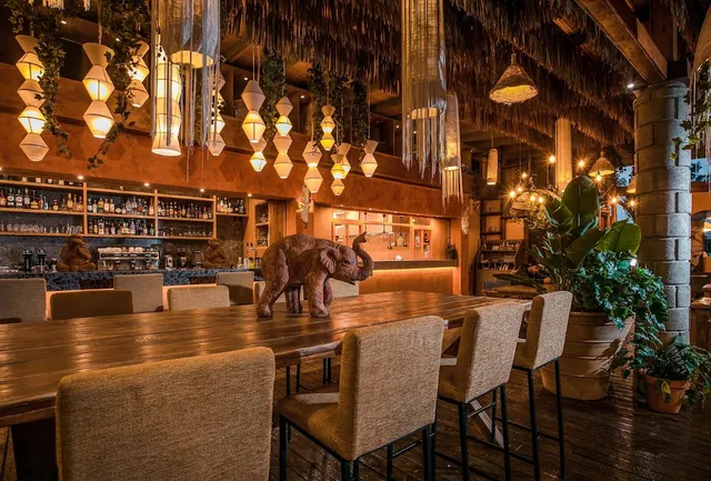 A long wooden table with bar stools, a wooden elephant sculpture and hanging lamps above