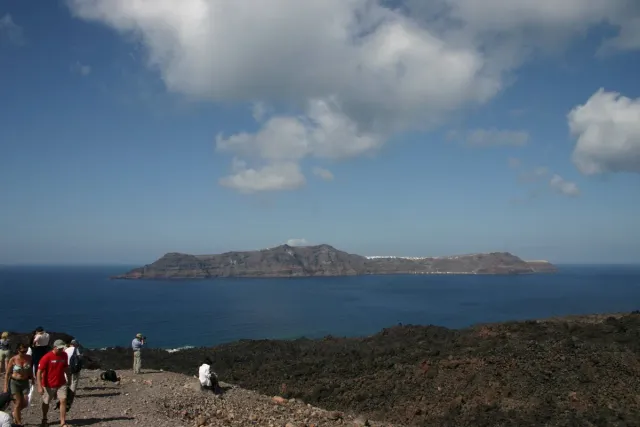 A hilly island across the blue caldera water, with walkers on a stony path in the foreground