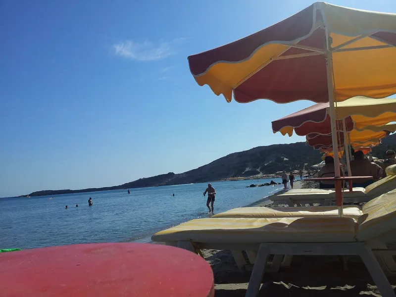 Sun beds and yellow umbrellas on Paradise beach in Kos, with bathers in the water and the bay slope behind