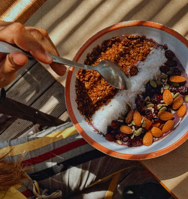 A hand with a spoon above a bowl of cereal, almonds, seeds and dried fruit in the sun