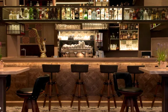 A bar counter with lit bottle shelves, tall stools and a saxophone on the wall