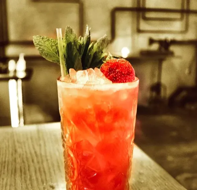 A red cocktail in a cut glass with ice, mint sprigs and a strawberry on the rim