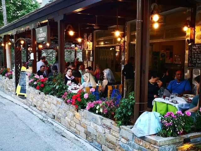 A covered terrace with diners, a stone wall and red geranium planters along the pavement