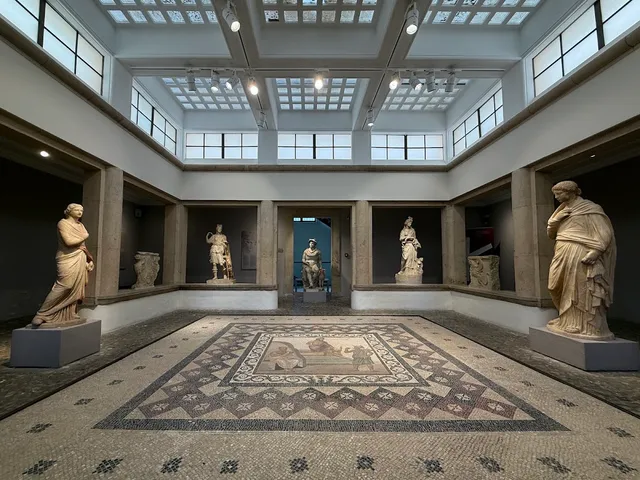 A hall with marble statues along the walls and a large mosaic in the center of the floor