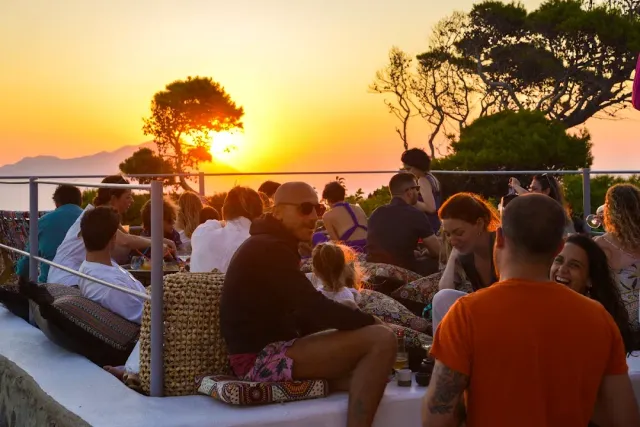 A crowd seated on cushions on a terrace at sunset, with pine trees and sea behind