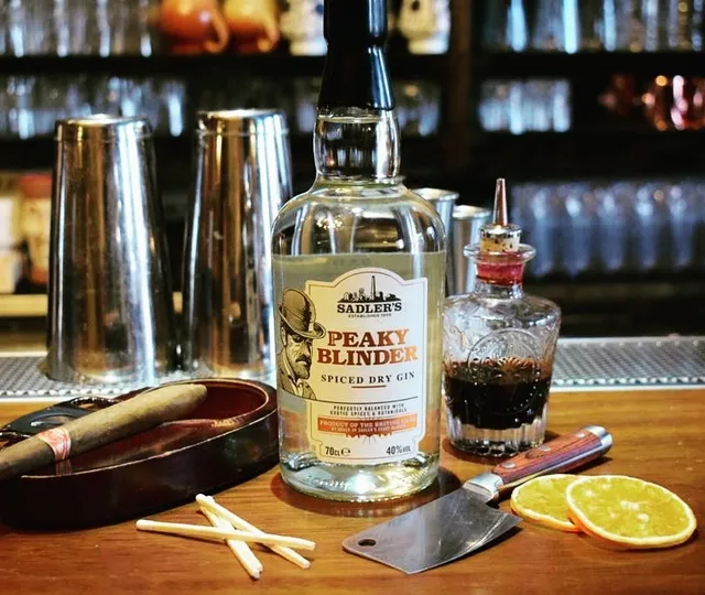 A gin bottle on a bar counter beside shaker tins, a cigar and orange slices