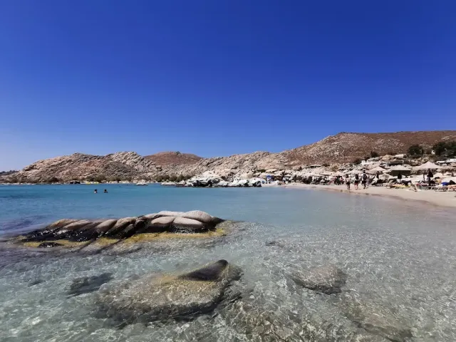 Shallow clear water with rounded rocks, and behind them a strip of sand and straw umbrellas