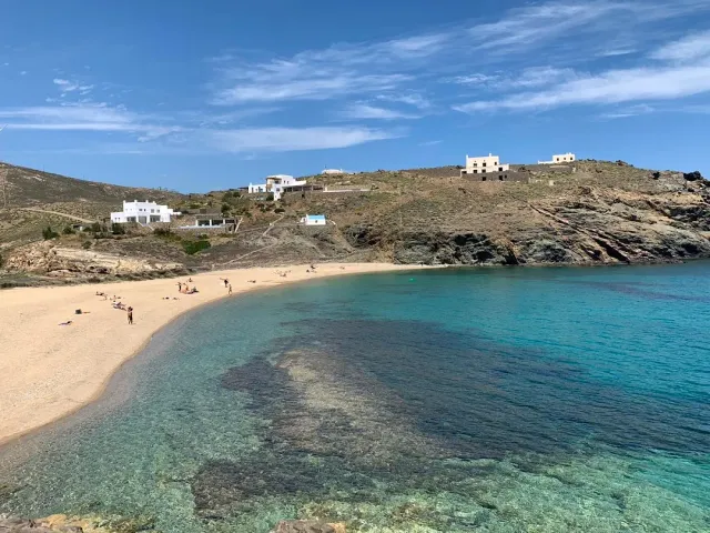 A small cove with pale sand, clear water and rocky patches, and a bare hill with white houses
