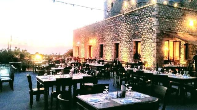 A terrace at dusk beside a lit stone building, dark laid tables and a string of bulbs