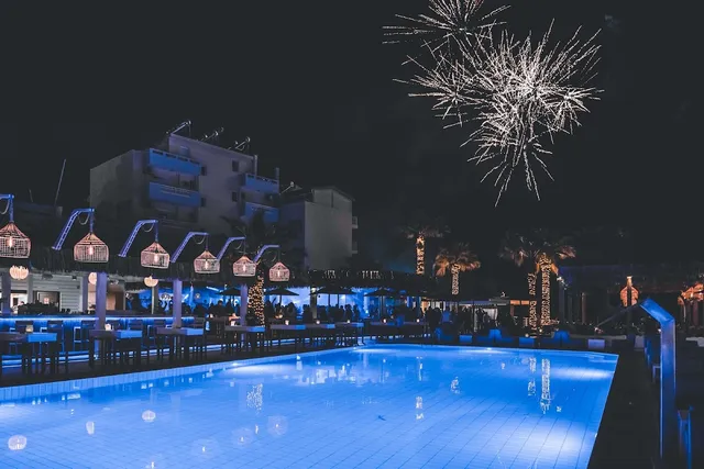 A pool lit in blue at night, with fireworks above the palm trees and tables