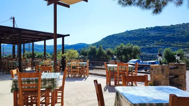 A terrace with a wooden pergola, checked tablecloths and chairs facing green hills and blue sky
