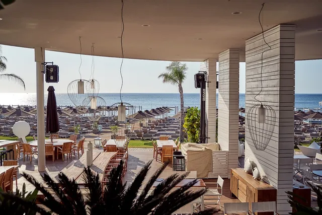 A covered terrace with wooden tables and wicker lamps, facing rows of parasols and the open sea