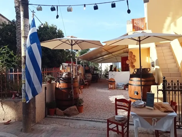 A yard with pale parasols, a hanging string of bulbs, wooden barrels and a Greek flag on a pole