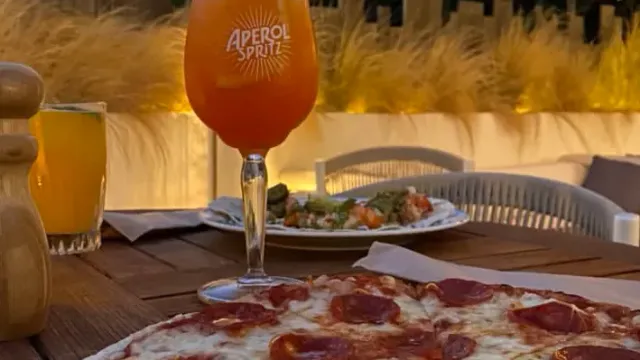 An orange cocktail glass on a wooden table, a pizza in front and another plate behind
