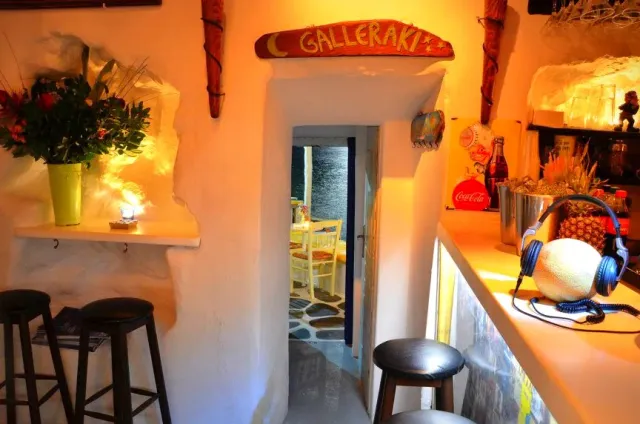 A narrow whitewashed passage in warm light, with bar stools, a wooden sign and an opening to a courtyard