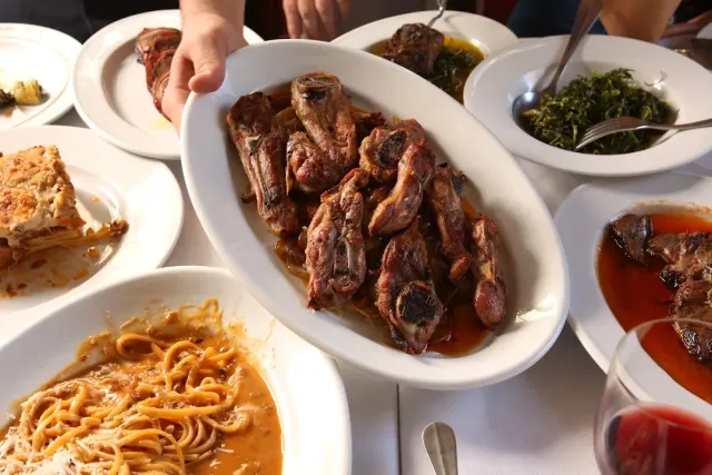 A hand serving a platter of grilled lamb chops, beside pasta in sauce and greens