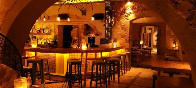 A vaulted stone space in orange lighting, with a lit counter and wooden stools