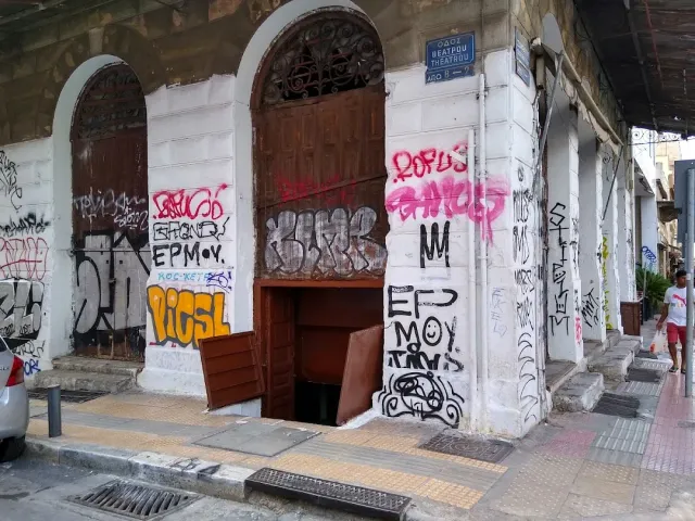 The corner of an old building covered in graffiti, with an open cellar hatch on the pavement