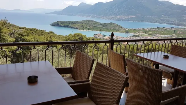 A balcony with wicker chairs and wooden tables above a green bay and mountains