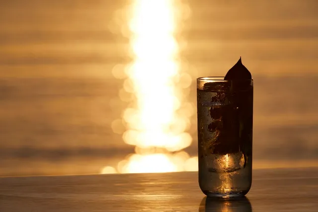 A glass of drink with ice on a counter, against golden sunset light reflected on the water