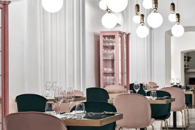 A dining room with pink and green armchairs, globe lamps and a pink glass cabinet