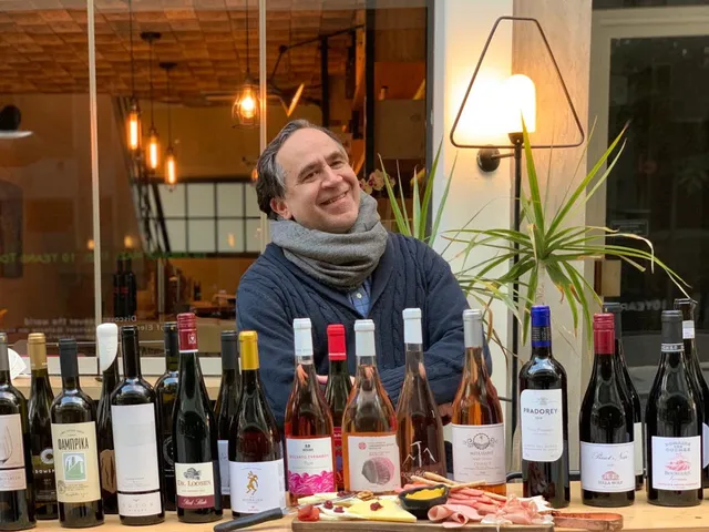 A smiling man behind a row of wine bottles and a platter of cured meats and cheeses