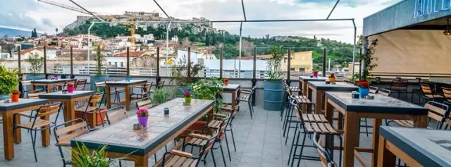 A rooftop terrace with wooden tables and chairs, and a hill with ancient ruins behind