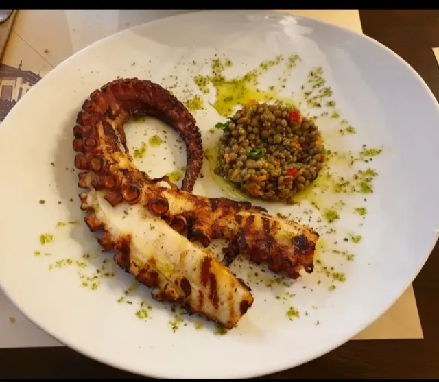 A grilled octopus arm on a white plate, beside a lentil salad and green oil