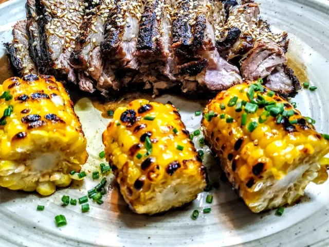 A sliced cut of meat with sesame seeds, beside three pieces of grilled corn