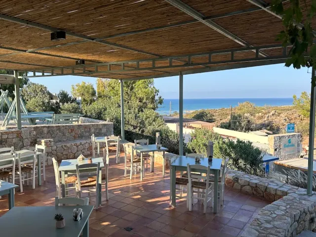A terrace under a reed canopy with pale blue tables and white chairs facing a strip of sea