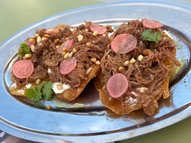 A metal platter with three fried discs topped with shredded meat, pine nuts, radish slices and coriander