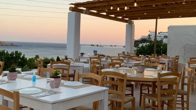 A terrace with a wooden pergola and bulbs, set tables and a pink sunset sky above the sea