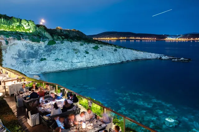 Diners on a clifftop terrace above a clear bay, under a blue evening sky