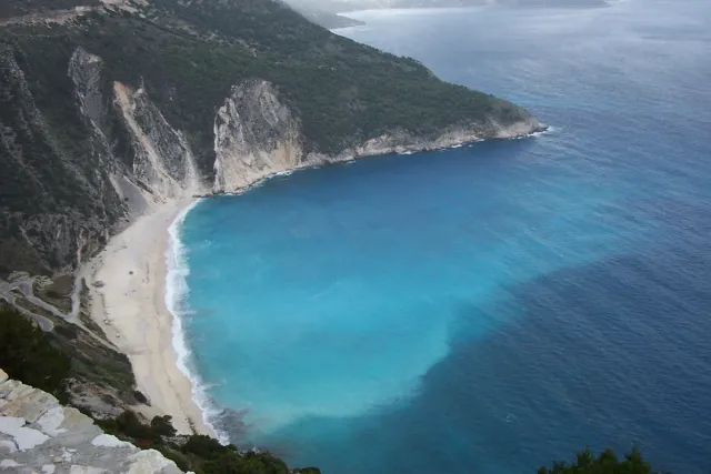 A bay with a white pebble shore and water in three shades of blue, between green cliffs