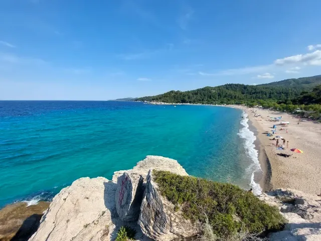 A turquoise bay and a long sandy beach below a wooded hill, seen from white rocks