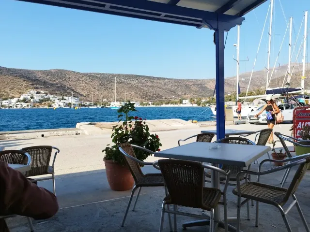 A terrace under a blue awning with tables and chairs facing a harbour with yachts and hills
