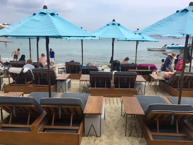 Blue parasols and wooden sunbeds on the sand facing the sea, with bathers in the background