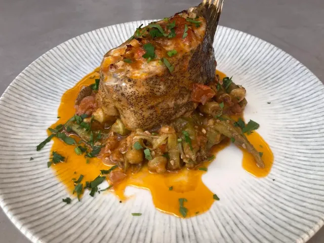 A cut of fish on a white plate over okra in tomato sauce, with chopped parsley