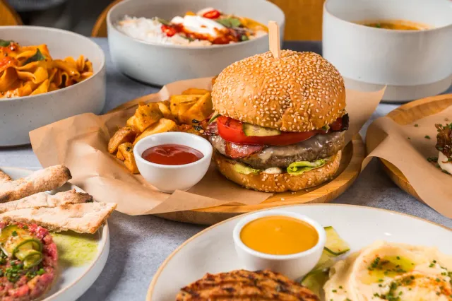A table loaded with dishes, and at its centre a burger in a sesame bun with potatoes