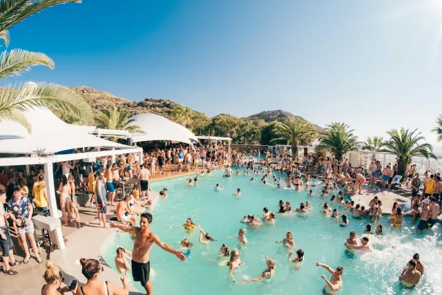 A large pool crowded with bathers, white canopies and palms around and a hill behind