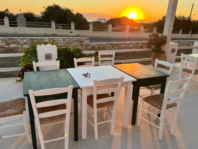 A terrace with simple tables and straw seat chairs facing a sunset over hills and a stone wall