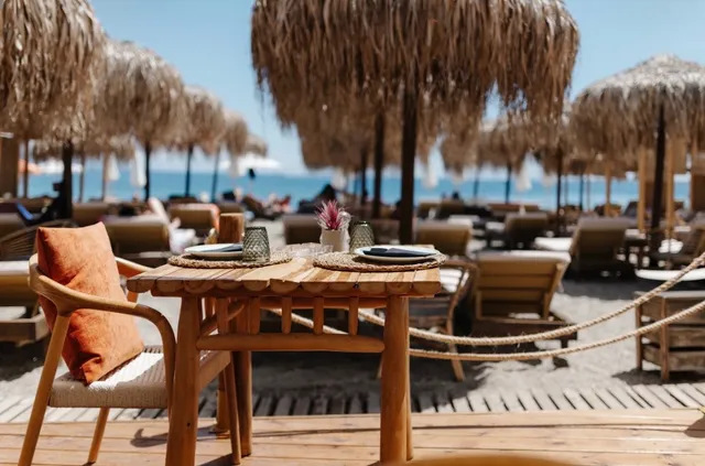 A wooden table set on a deck facing the beach, with straw umbrellas and sunbeds down to the sea