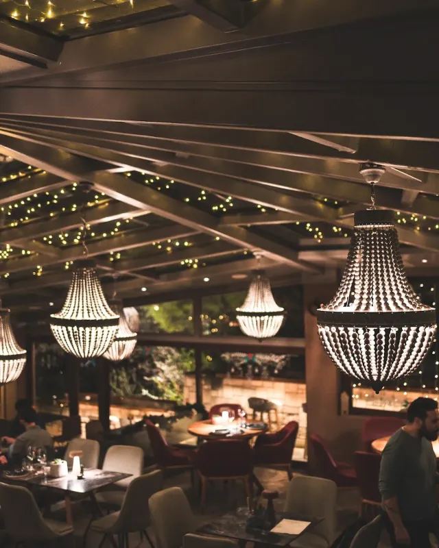 A covered space in the evening with beaded chandeliers, strings of lights and armchairs around tables