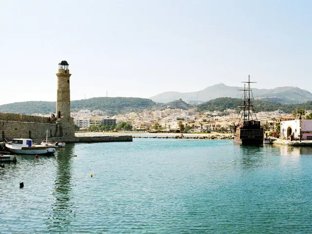 A stone lighthouse at the end of a pier, with a harbour of boats and the town behind