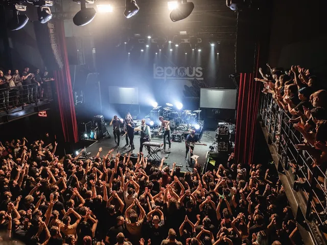 A full concert hall seen from above, a cheering crowd before a stage flooded with white lights