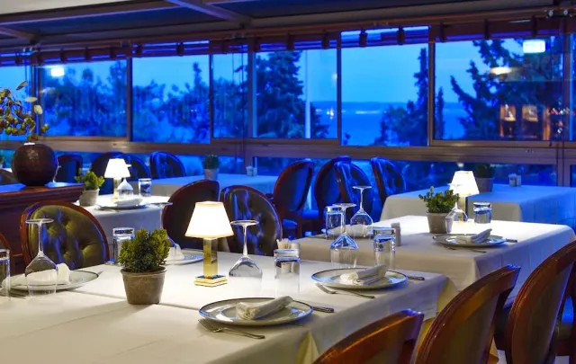 A dining room with white cloths and small table lamps, facing windows at blue hour