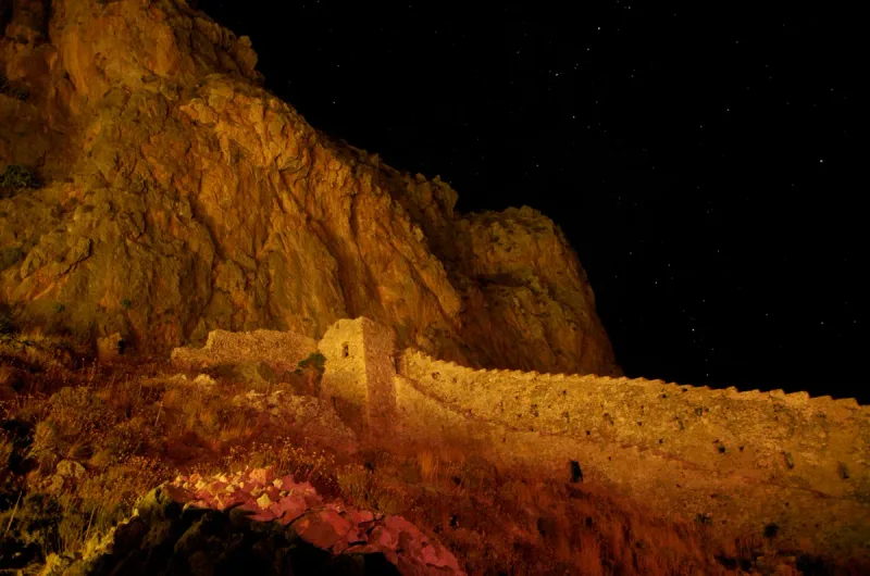 The fortress wall of Monemvasia floodlit at night at the foot of the great rock, under a starry sky