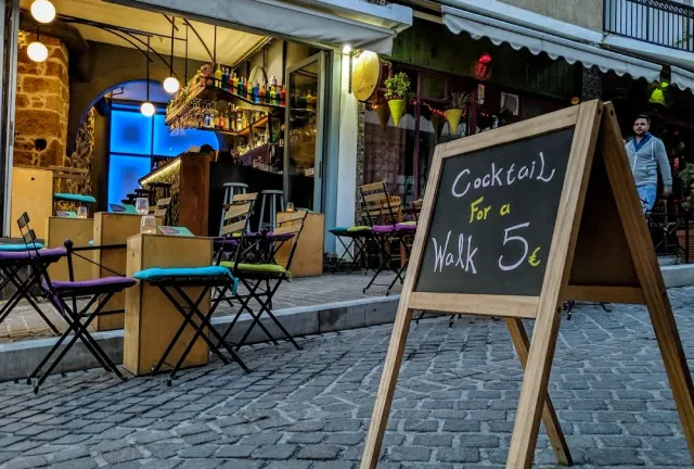 A paved alley in the evening with colourful tables, a lit bar front and a chalkboard