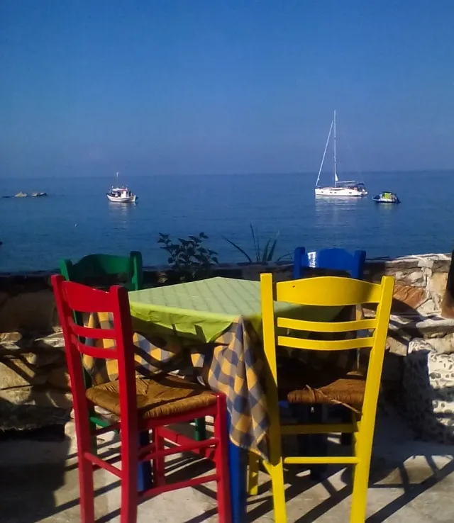 Colourful chairs around a table with a green cloth facing a blue sea with a sailing boat